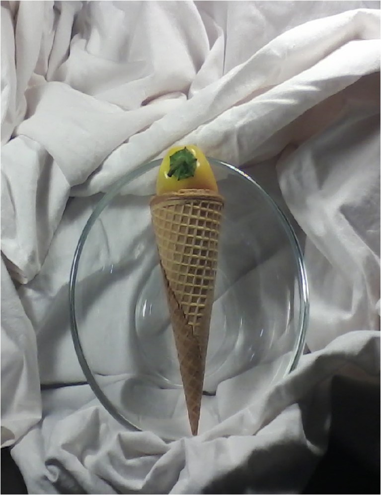 Clear glass bowl with yellow pepper inside of a sugar waffle ice cream cone standing in center, with white fabric surrounding
